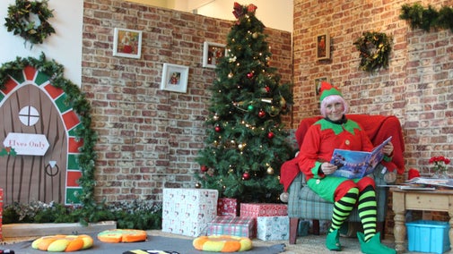 A photograph of an person dressed up as an elf sitting on a chair holding a book. The room is decorated in a Christmas theme with a Christmas tree in the centre of the image.